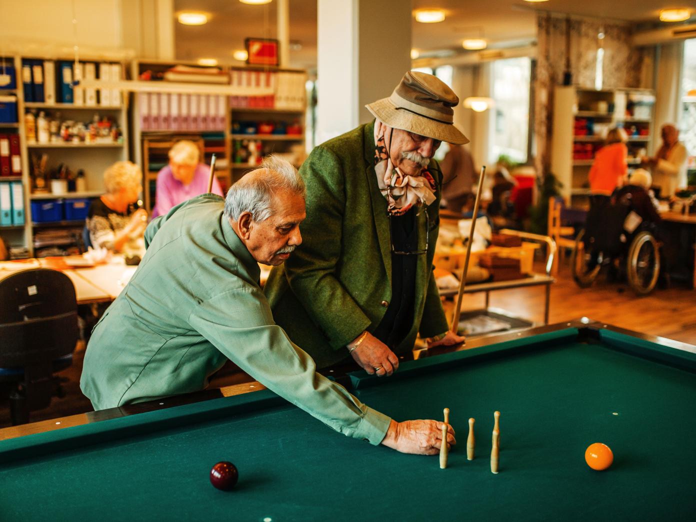 To ældre herrer med anden etnisk baggrund end dansk sætter kegler op på et billardbord. I baggrunden sidder der mennesker og taler omkring borde.