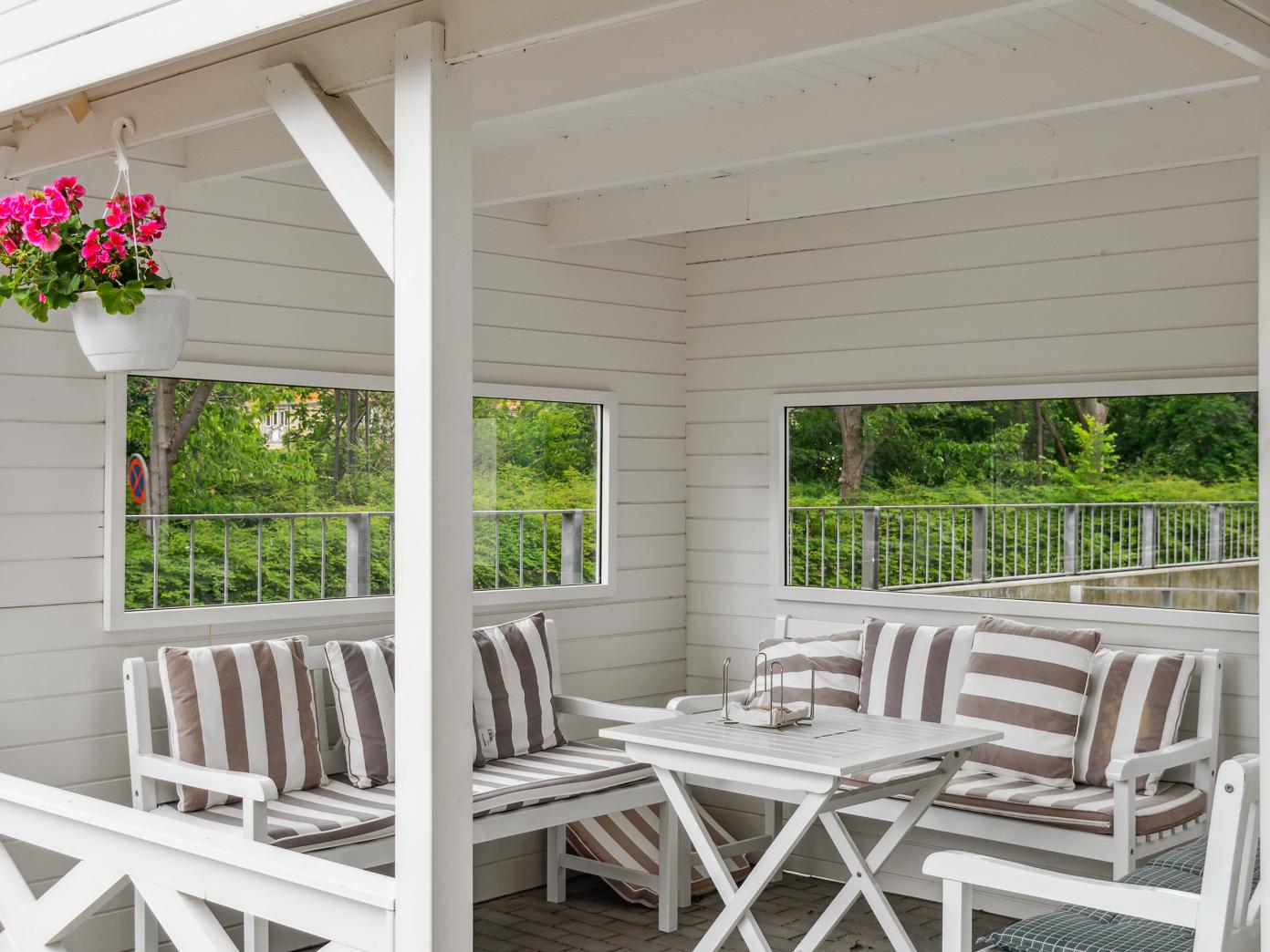Overdækket terrasse i hvidmalet træ med bænke, borde, stole og hynder. En lyserød blomster hænger fra en bjælke.