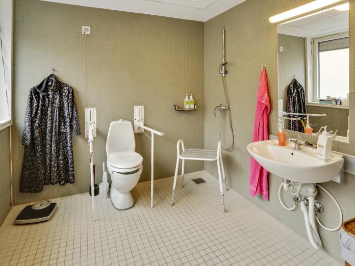 Badeværelse med handicapfaciliteter, badebænk, vask, spejl. På væggen hænger en jakke, og på gulvet står en badevægt. 
