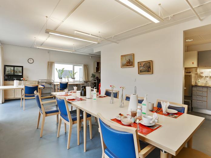 Fællesrum med langt spisebord med seks stole, kig til køkken og flere møbler i baggrunden.