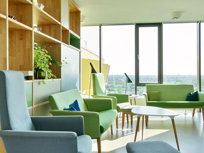 Fælles stue i rum med gulv til loft-vinduer. Stuen er indrettet med reol, sofaer, lænestole, sofabord og standerlamper samt grønne planter i krukker.