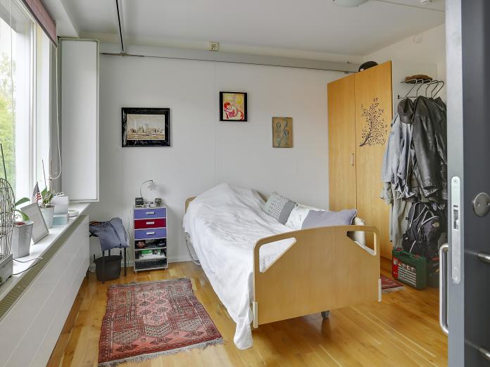 Soveværelse med plejeseng, skab med to høje låger, sengemøbel og et tæppe på gulvet. I loftet er der skinner til loftlift. 