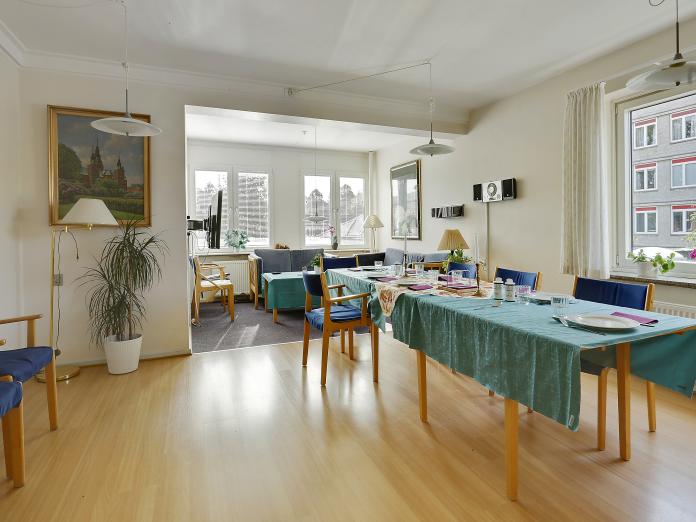 Fælles stue med langt spisebord og stole samt hyggekrog med sofaer, lænestole og sofabord samt grønne planter, standerlamper og malerier på væggene.