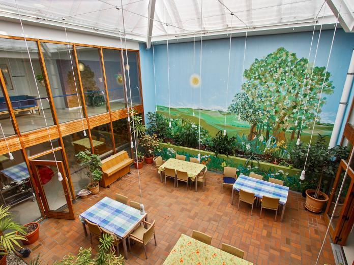 Overdækket atriumgård med glasfacader og vægmaleri på den ene væg. Rummet er indrettet med borde, stole, klaver og mange grønne planter i krukker.