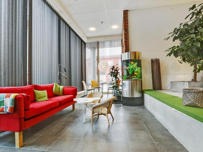 Moderne fællesområde med meget højt til loftet og en væg af vinduer ud mod gaden. Rummet indeholder grønne planter, et akvarie, en sofa og flere områder med siddepladser.