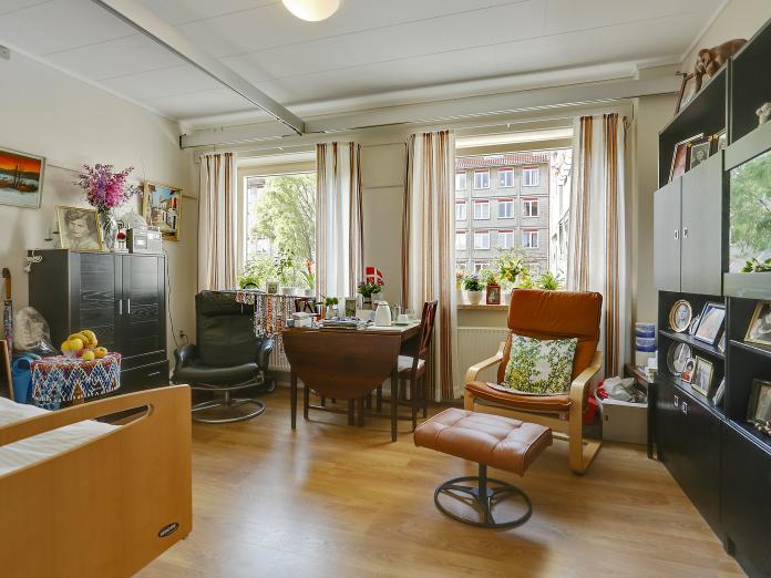 Lys stue med lyse gulve og to vinduer ud mod fælles have. Stuen er møbleret med en plejeseng, en loftslift og personlige møbler som lænestole og spisebord med stole.