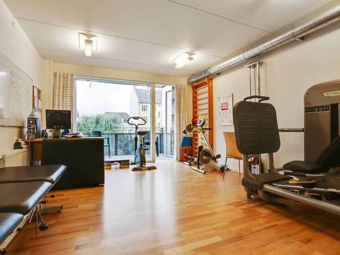 Motionsrum med kondicykel, siddende kondicykel, ribbe, briks og andre træningsmaskiner. Rummet er lyst med udgang til altan. 