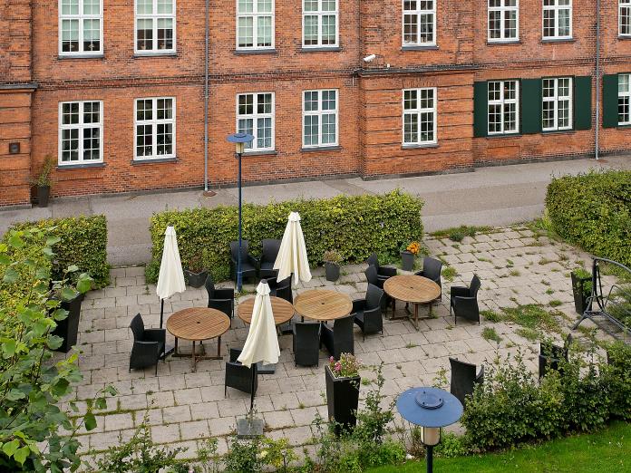 Fliseterrasse omkranset af grønne hække. Terrassen er indrettet med runde borde, kurvestole og parasoller samt blomster i krukker.