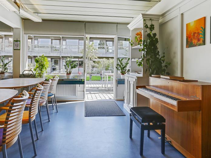 Fællesrum med store vinduespartier og en åben dør ud mod fælles terrasse. Rummet er møbleret med moderne borde og stole, klaver og grønne planter i vindueskarmene.
