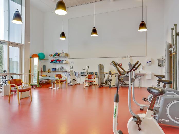 Stort motionsrum med højt til loftet og store vinduespartier. Rummet indeholder mange træningsmaskiner som kondicykler samt behandlingsbriks. 