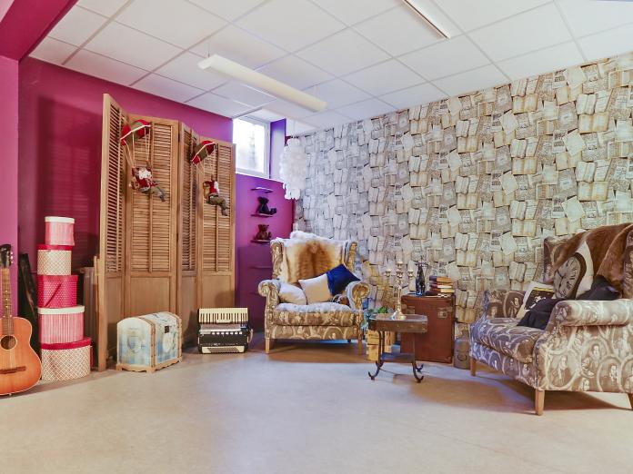 Hyggekrog med væg tapetseret med gamle bøger og indrettet med bløde lænestole, en gammel kuffert, en guitar og andre stemningsfulde genstand.