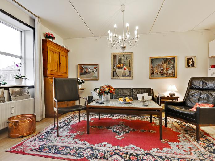 Lys stue indrettet med personlige møbler som sofa, lænestole, sofabord samt chatol. På væggene hænger malerier og i loftet en lysekrone.