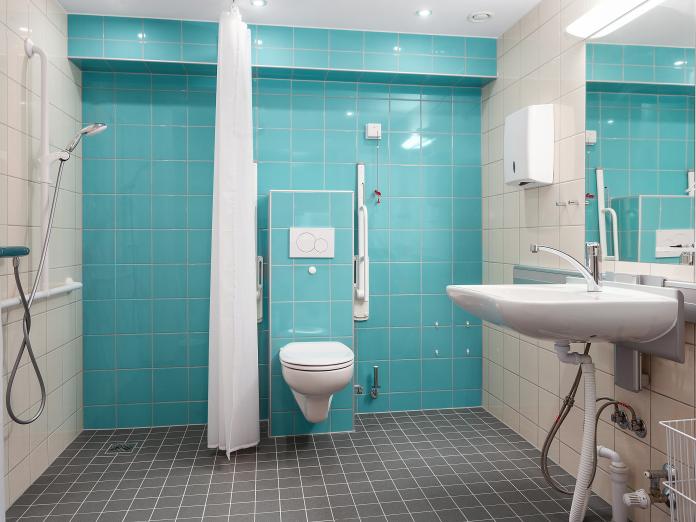 Stort badeværelse i hvide, grå og turkise fliser med bruser, væghængt toilet, vaske og spejl.