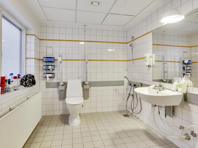 Stort, lyst badeværelse i hvide klinker med toilet, vask, stort spejl og bruser.  