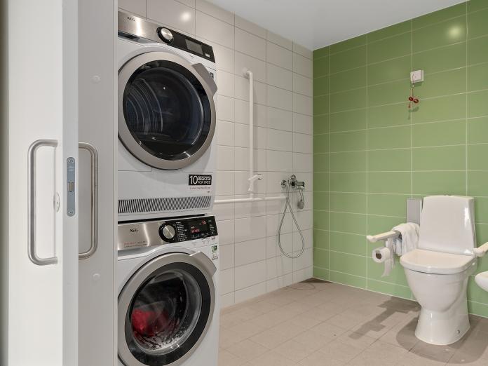 Stort badeværelse i hvide og grønne klinker med toilet, håndvaske og bruser samt vaskesøjle bestående af vaskemaskine og tørretumbler.