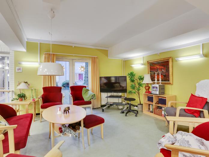 Fælles stue med lysegrønne vægge indrettet med lænestole, sofa og sofabord samt fjernsyn, grønne planter og nipsgenstande. 