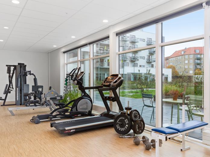 Motionsrum med store vinduespartier ud til gård indrettet med forskellige træningsmaskiner som løbebånd, romaskine og frivægte.