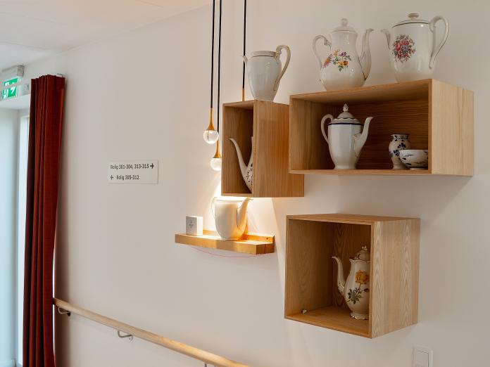 Samling af træbogkasser på hvid væg. I bogkasserne ses kaffekander fra forskellige porcelænsstel.  