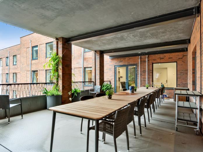 Fælles, overdækket terrasse med langbord, fletstole, potteplanter på bordet samt store krukker med grønne planter.