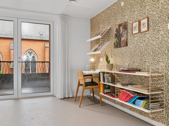 Hjørne af fælles stue med tapetseret væg, hvorpå der hænger en string reol med skrivebord og hylder med bøger og potteplante samt billeder på væggen.  