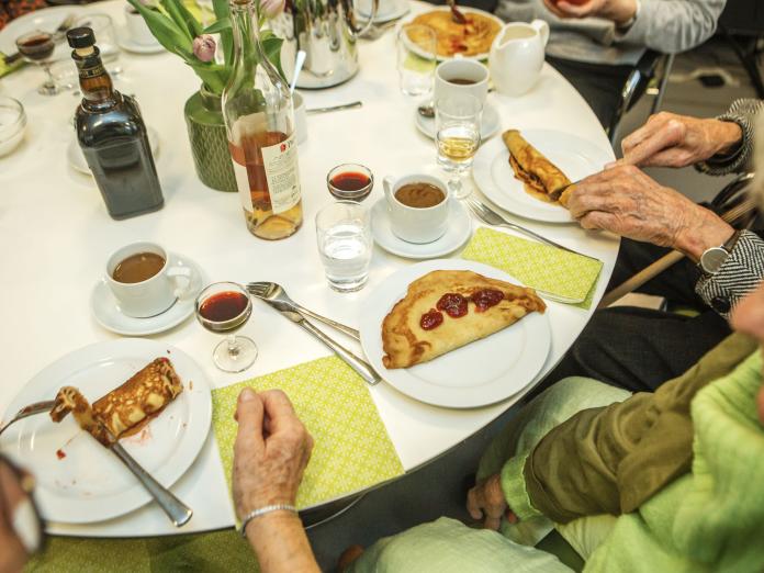 Ældre personer sidder om et rundt bord og spiser pandekager med syltetøj. På bordet er der kaffe, portvin og cognac samt eb buket blomster i en vase.