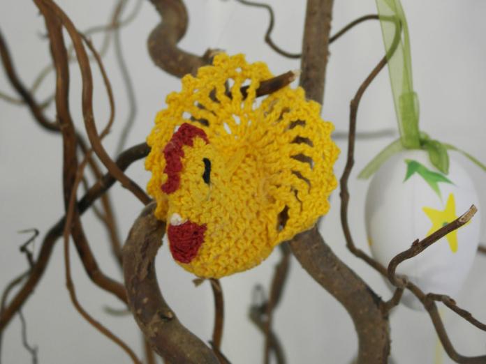 En lille, gul hæklet kylling, der hænger på troldegrene og et påskeæg af porcelæn.