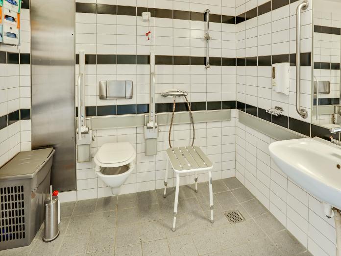 Lyst flisebadeværelse med vask, spejl, toilet med armstøtter, bruser og badebænk samt indbygget skab.