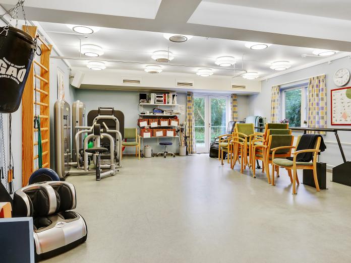 Stort, lyst motionsrum indrettet med træningsmaskiner, en ribbe, gangbarre, massagestol og stole til siddende træning.