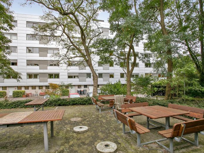 Aftensols grå bygninger set fra terrasse med borde-bænke-sæt og høje træer.