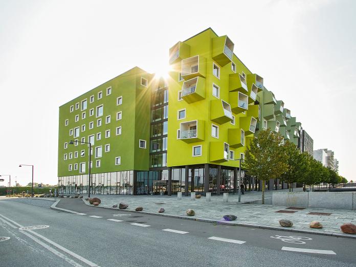 Moderne bygning i grønne og gule farver med altaner og hvide vinduer. Foran bygningen er en stor, flisebelagt plads med træer og en vej.