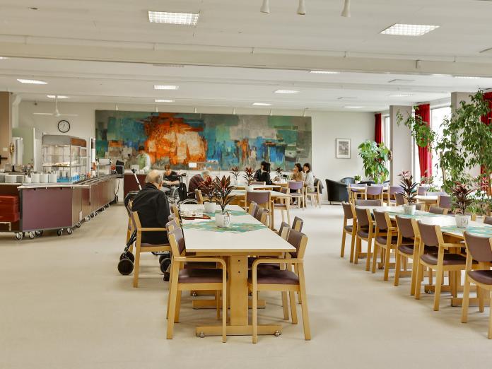 Fælles spisestue med langborde og stole, grønne planter og kunst på endevæggen.
