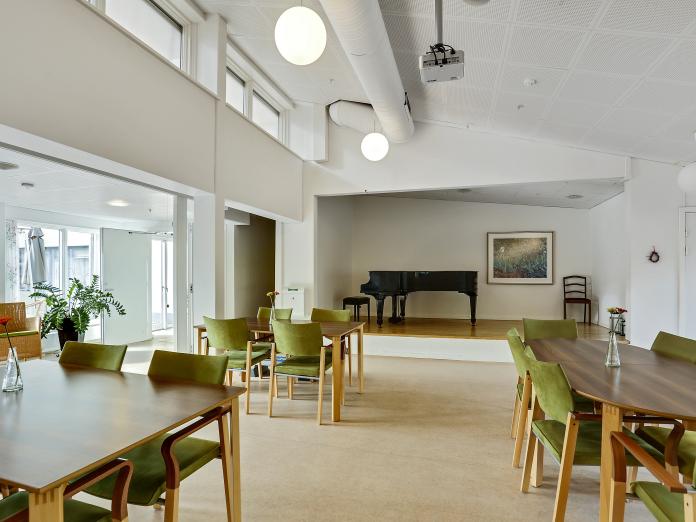 Fællesrum med borde, grønne stole, højt til loftet og vinduer og et stort klaver, der står hævet en smule op på en lille scene.