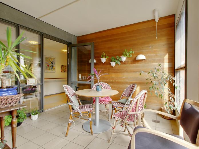 Rum indrettet som terrasse med træbeklædte vægge, fliser på gulvet og masser af grønne planter i krukker og på væggen. Rummet er indrettet med et cafébord og kurvestole.