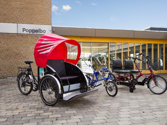 Tre forskellige slags cykler parkeret på fliseterrasse. Der er både en rickshaw, som er en moderne ladcykel til persontransport, og to duocykler. 