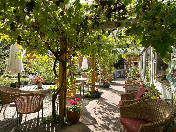 Overdækket terrasse med vinrænker, kurvemøbler og blomster i krukker. 