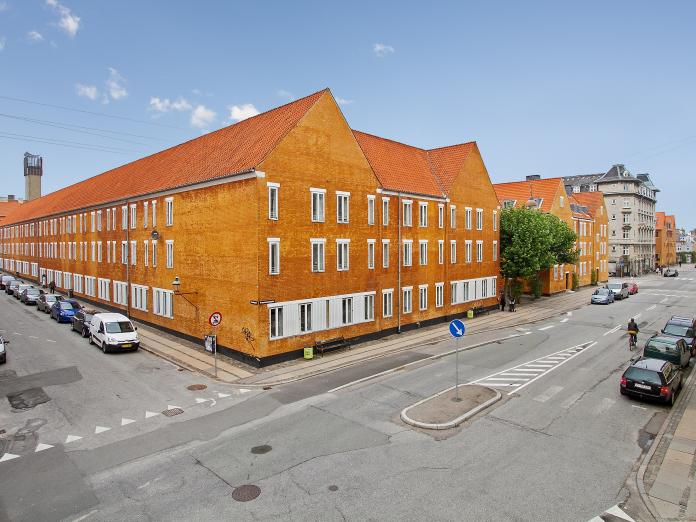 Stort gulkalket murstensbyggeri med hvide vinduer og røde tegl. Bygningen er omkranset af vej, hvor der holder biler parkeret.