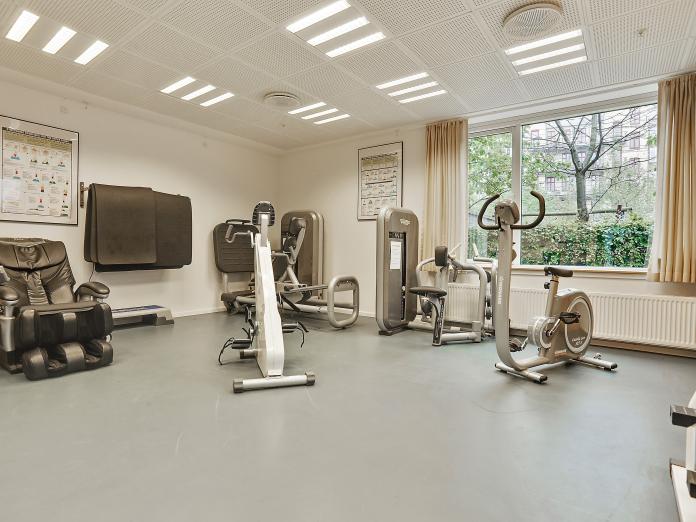 Motionsrum med forskellige maskiner, bl.a. en cykel og massagestol. På væggen hænger yogamåtter og plakater.