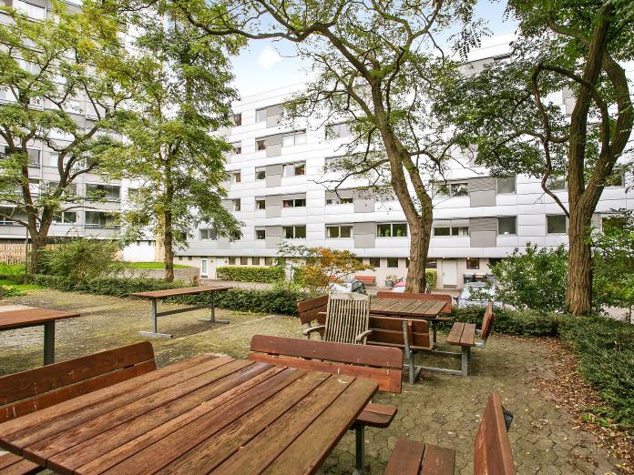 Aftensols grå bygninger set fra terrasse med borde-bænke-sæt og høje træer.