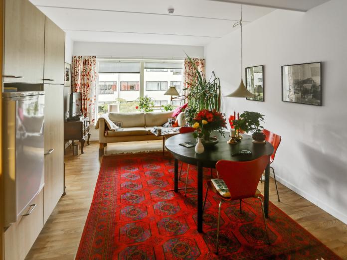 Stue med tekøkken, tæppe, lille rundt spisebord med røde stole, sofa, sofabord og tv-møbel stående foran et stort vinduesparti.