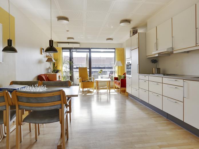 Fællesområde med større køkken, sofaarrangement, spisebord og stole samt gulv til loft-vinduer ud til overdækket altan.