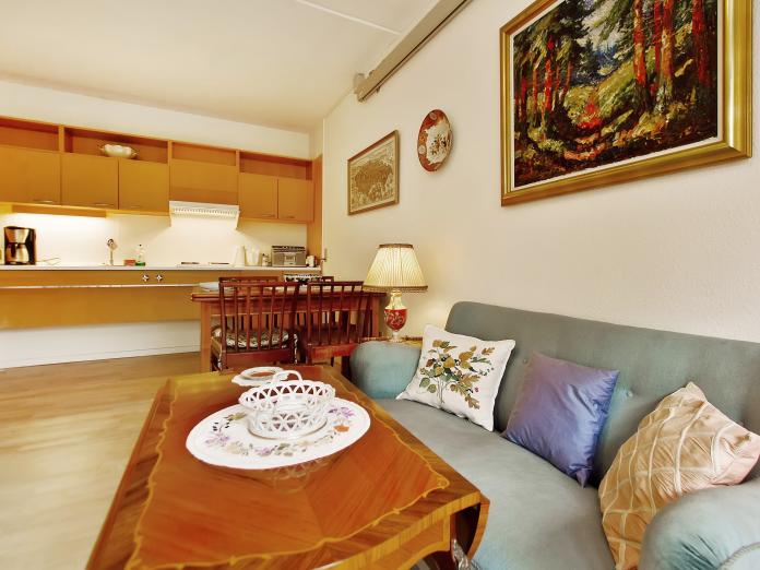 Stue med hvidmalede vægge, lyse gulve, sofa, spiseplads med bord og stole samt køkken i lyst træ. 