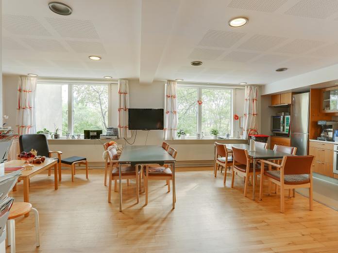 Fællesrum med borde, køkken, lænestol, sofabord, tv på væggen og to store vinduespartier.