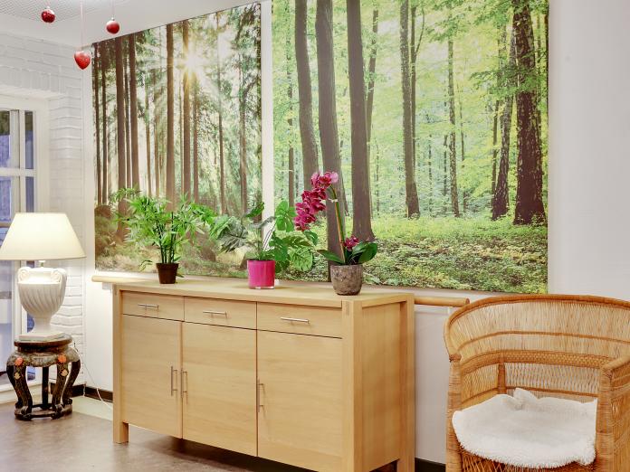 Hjørne dekoreret med fotostater af skov på væggen og indrettet med skænk, kurvestol og et sidebord med lampe.