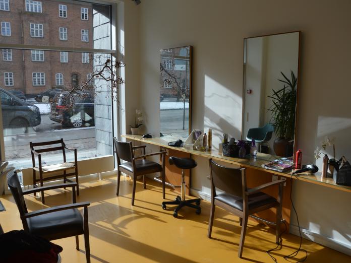 Frisørsalon med langt bord med hårprodukter og hårtørrer samt fem stole. Der er store spejle på væggen og et stort vindue med udsigt ud til gaden. 