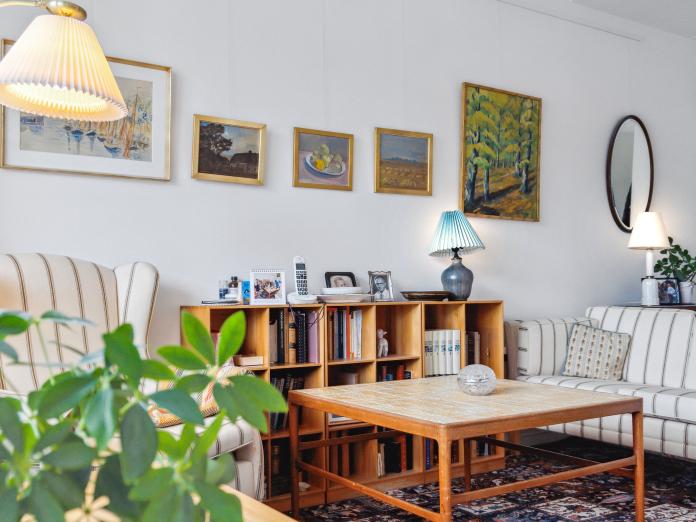 En bolig med sofa, stol, billeder på væggen, en lampe, en lav bogreol og grønne planter.