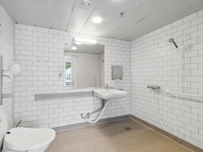 Stort, handicapvenligt badeværelse med hvide fliser på væggene, toilet, vask og åben bruseniche.