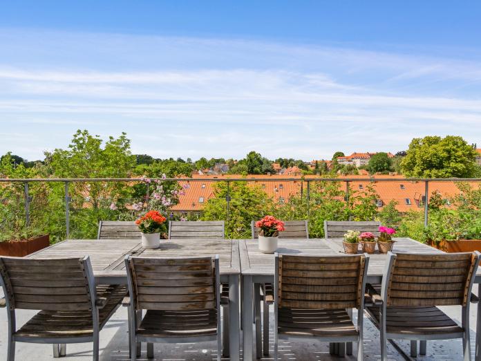 Fælles terrasse med udendørsmøblement, blomster på bordene og udsigt til træer og masser af røde tage.