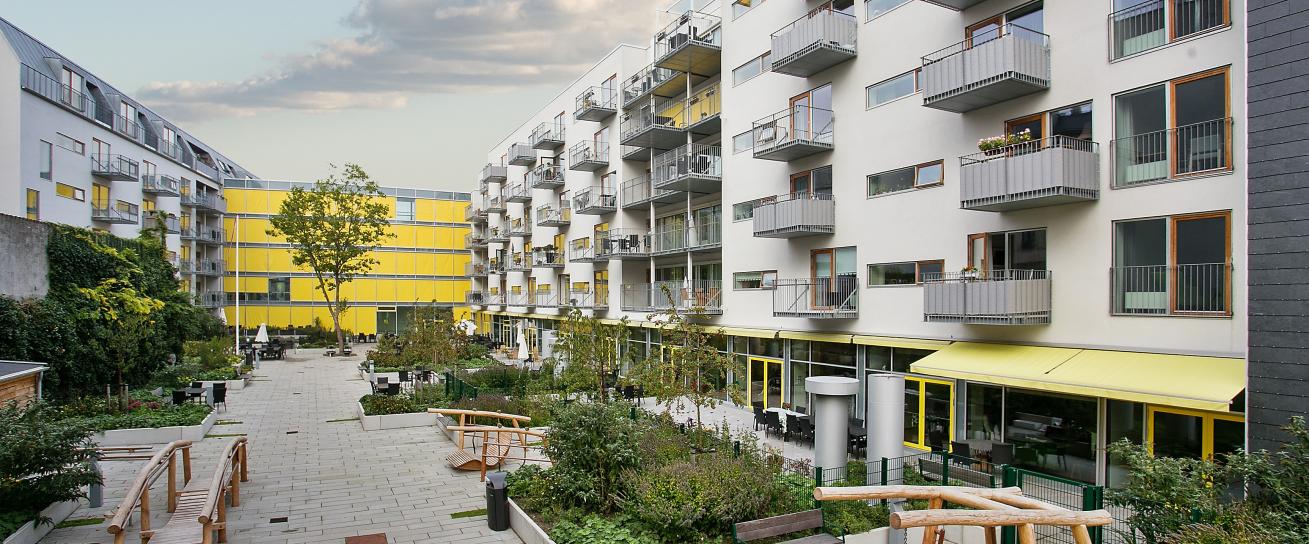 Moderne bygninger i hvide og gule farver med hvide og træfarvede vinduer samt stålaltaner og ståltag. Mellem bygningerne ses en flisebelagt gårdhave med bede, træer og havemøbler.